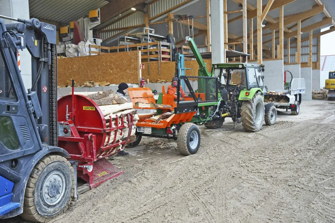 Ein Gabelstapler lädt Holz auf einen Tischholzspalter. Ein Traktor mit angehängtem Wagen wartet daneben.
