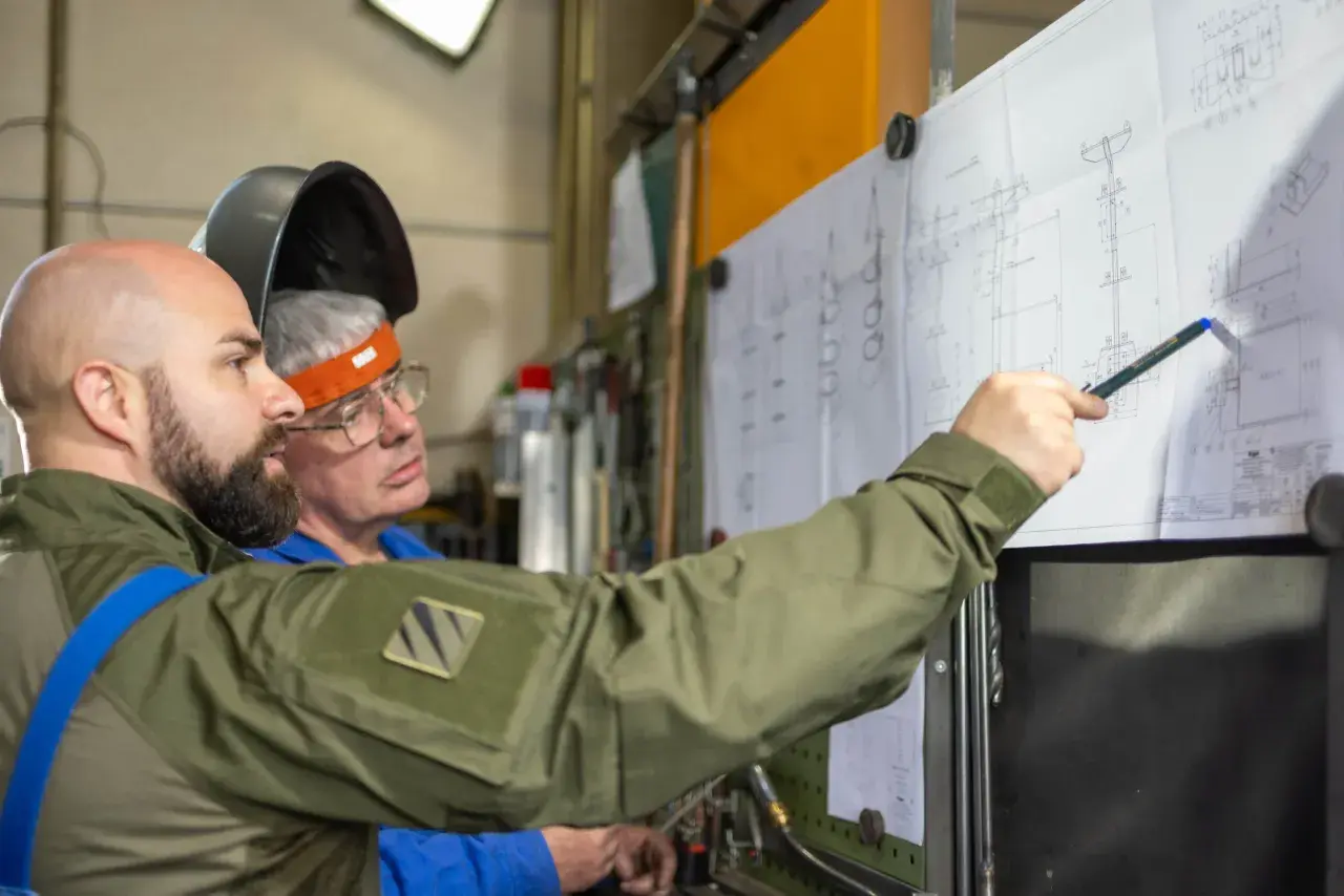 Zwei Männer besprechen einen technischen Plan. Einer zeigt mit einem Stift auf Details, die an einen **Ratschenzug** erinnern.