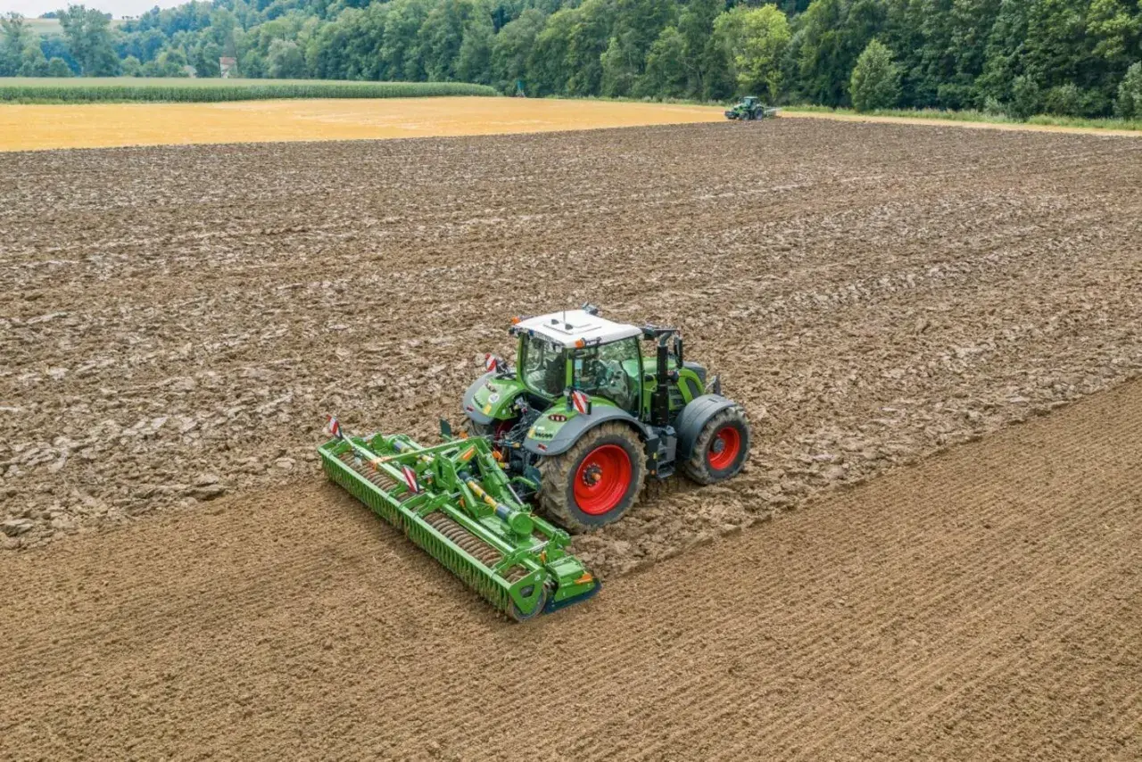 Grüner Traktor mit einem breiten kreiselgrubber bearbeitet ein feuchtes Feld. Im Hintergrund ein weiteres Feld und Wald.
