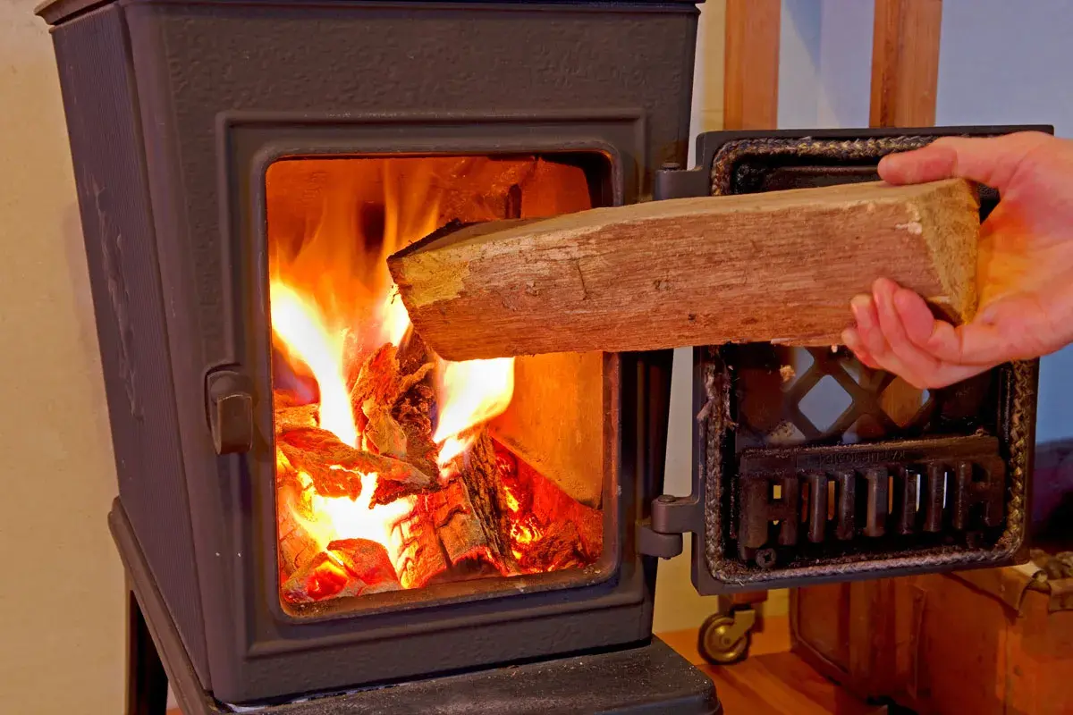 Hand legt Holzscheit in brennenden Ofen. Achtung: welches Holz darf man nicht verbrennen?