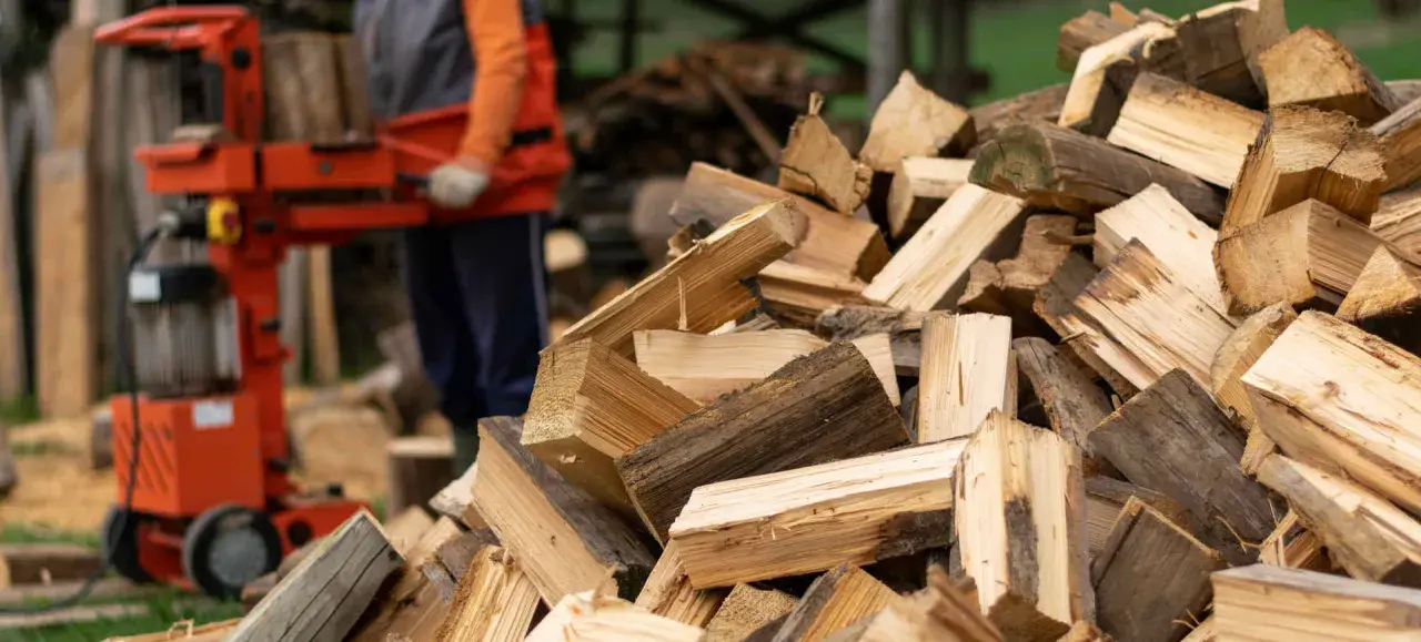 Ein Mann bedient einen roten Holtzspalter, während ein großer Haufen gespaltenes Holz daneben liegt.