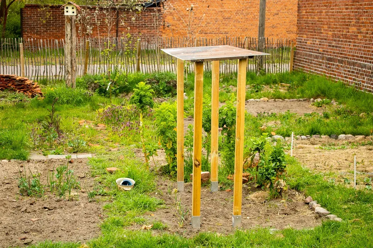 Ein provisorisches tomatendach selber bauen: Holzpfosten mit Metallfüßen tragen eine einfache Dachkonstruktion im Garten.