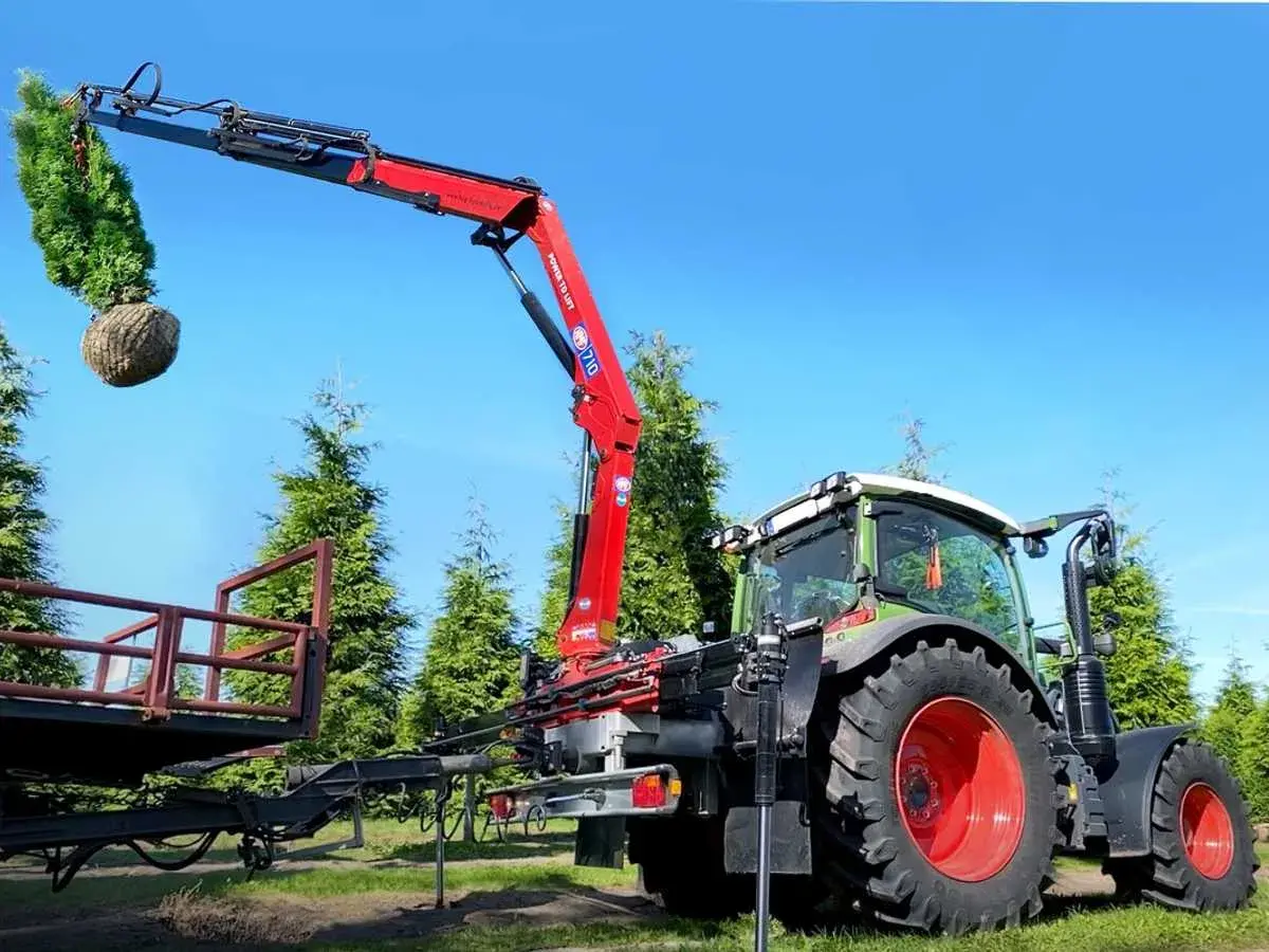 Ein roter Ladekran, gebraucht für Traktor, hebt einen Baum mit Wurzelballen. Der Traktor steht daneben, bereit für den Transport.