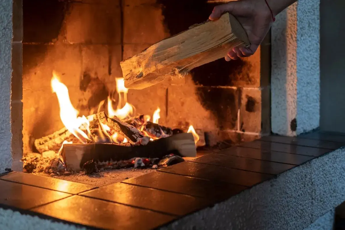 Eine Hand legt Holz nach, das im Kaminfeuer brennt. Das Holz brennt nicht, es spendet Wärme.