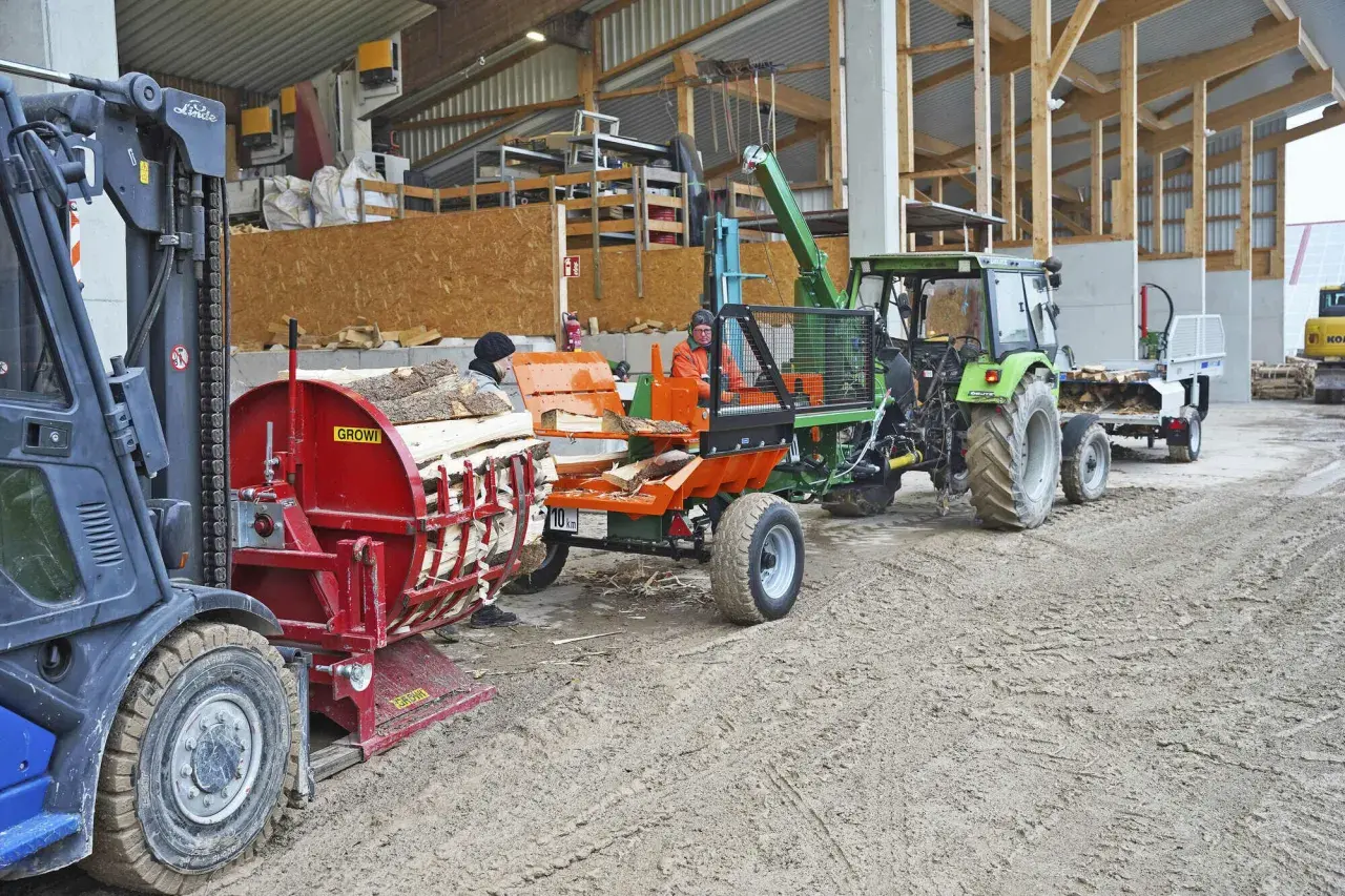 Gabelstapler lädt Holz auf eine hydraulische Kreissäge. Ein Traktor mit Anhänger wartet daneben.