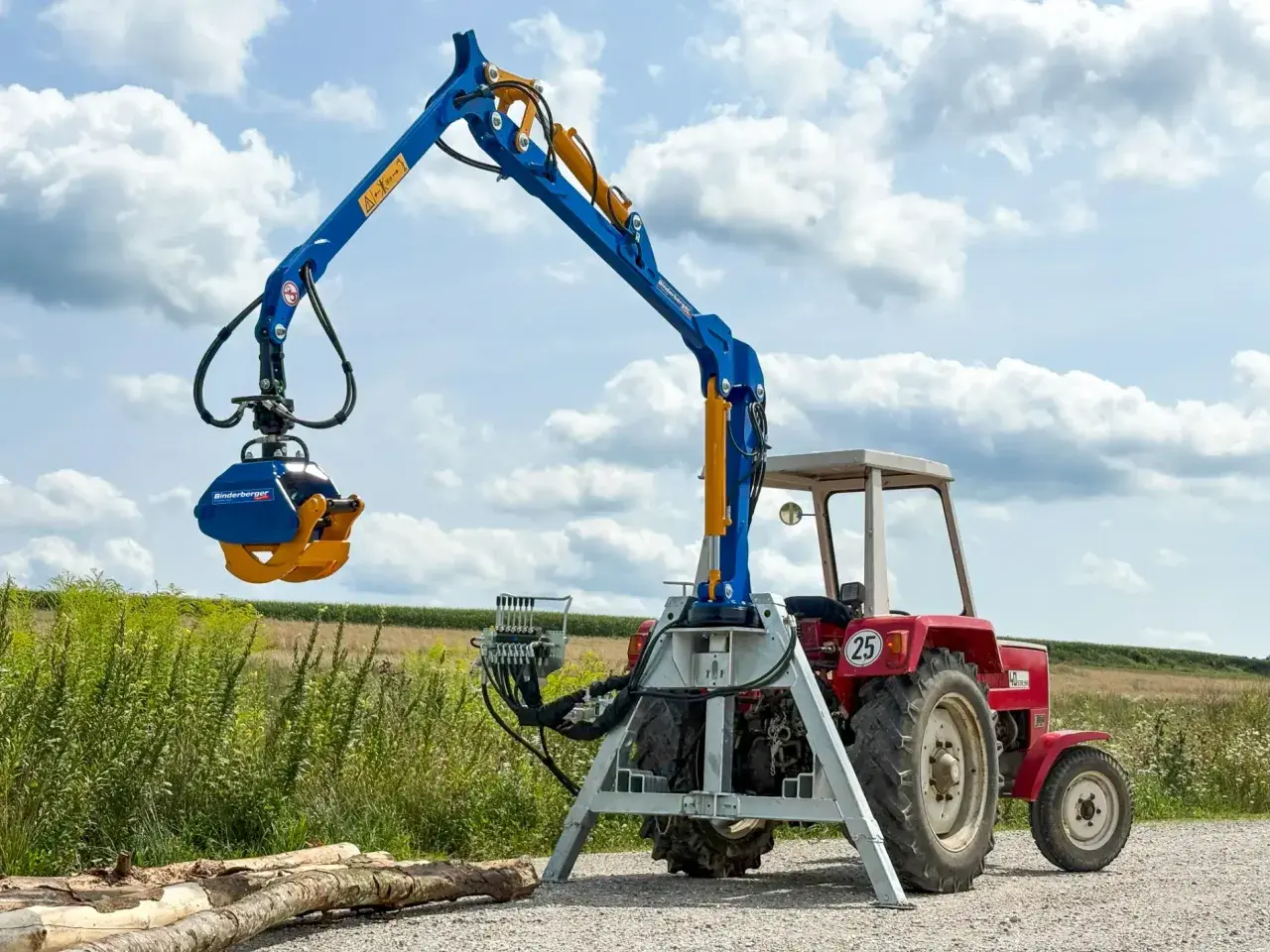 Ein blauer Forstkran mit Dreipunkt-Aufnahme und Greifer steht neben einem roten Traktor im Feld.