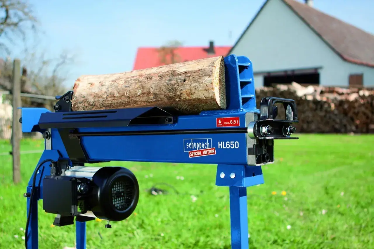 Ein liegender bgu Holzspalter HL650 von Scheppach mit einem Holzstamm bereit zum Spalten. Im Hintergrund ein Haus und Holzstapel.
