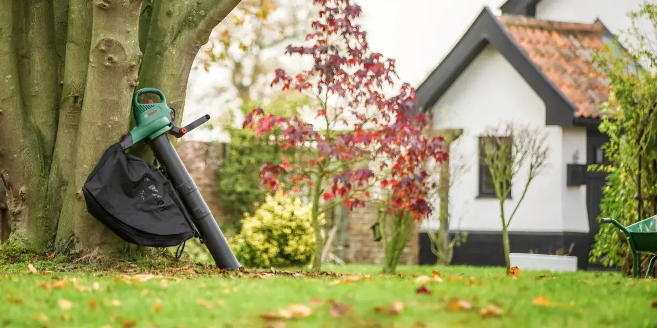 Laubbläser mit Fangbeutel lehnt am Baum, bereit, das Herbstlaub zu zerkleinern. Im Hintergrund ein Haus und Garten.