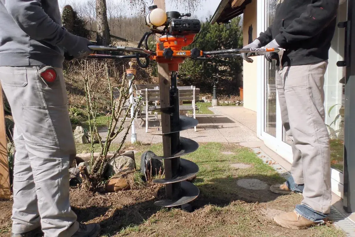 Zwei Personen bedienen einen leistungsstarken **Erdhandbohrer** im Garten. Das Gerät mit seinem orangefarbenen Motor und der langen Bohrschnecke ist bereit, Löcher für Pfosten oder Pflanzen zu graben.