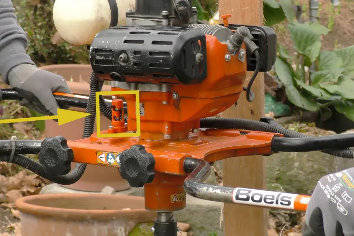 Ein orangefarbener Erdbohrer im Garten, bereit für den Einsatz. Ein gelber Pfeil hebt einen roten Hebel hervor.