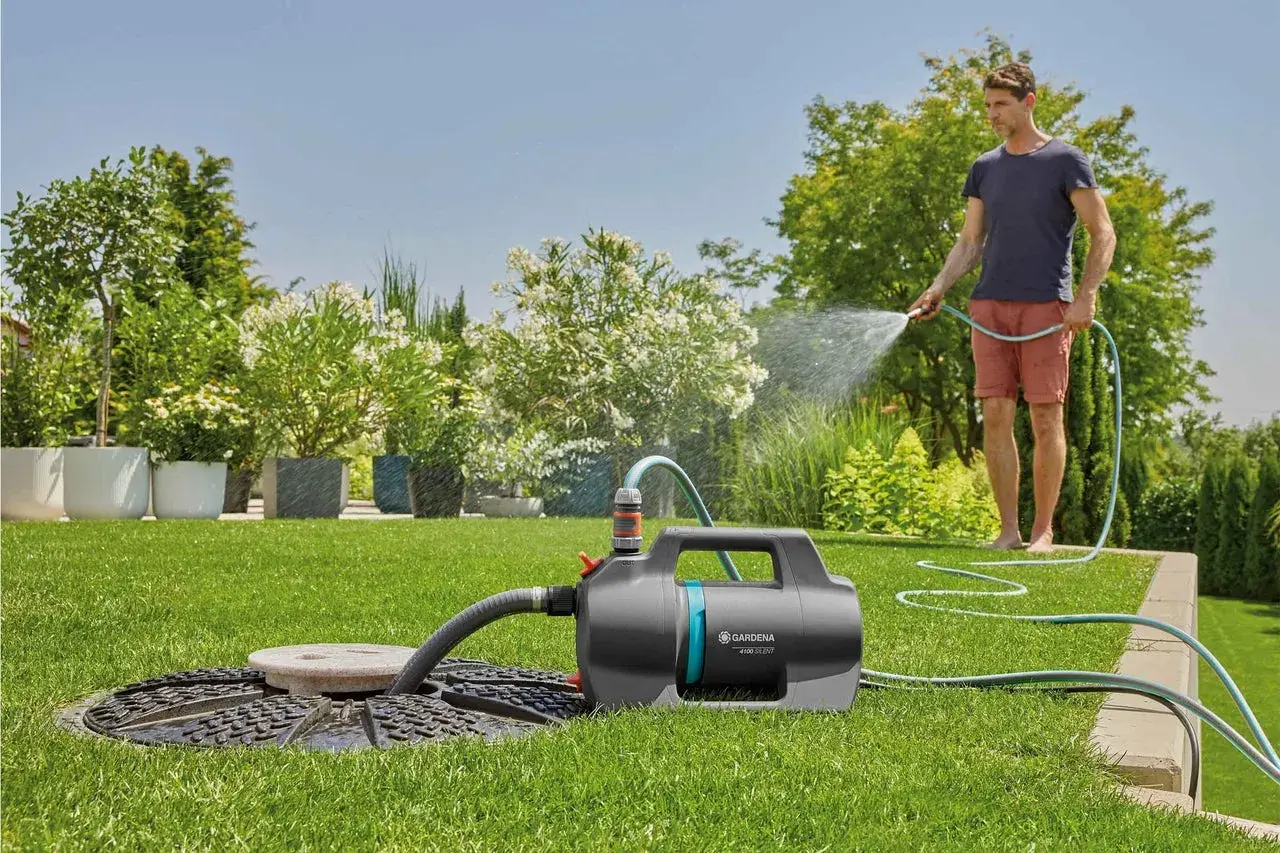 Mann bewässert den Garten mit einer Tauchpumpe, die aus einem Schacht saugt. Üppige Pflanzen und ein gepflegter Rasen.