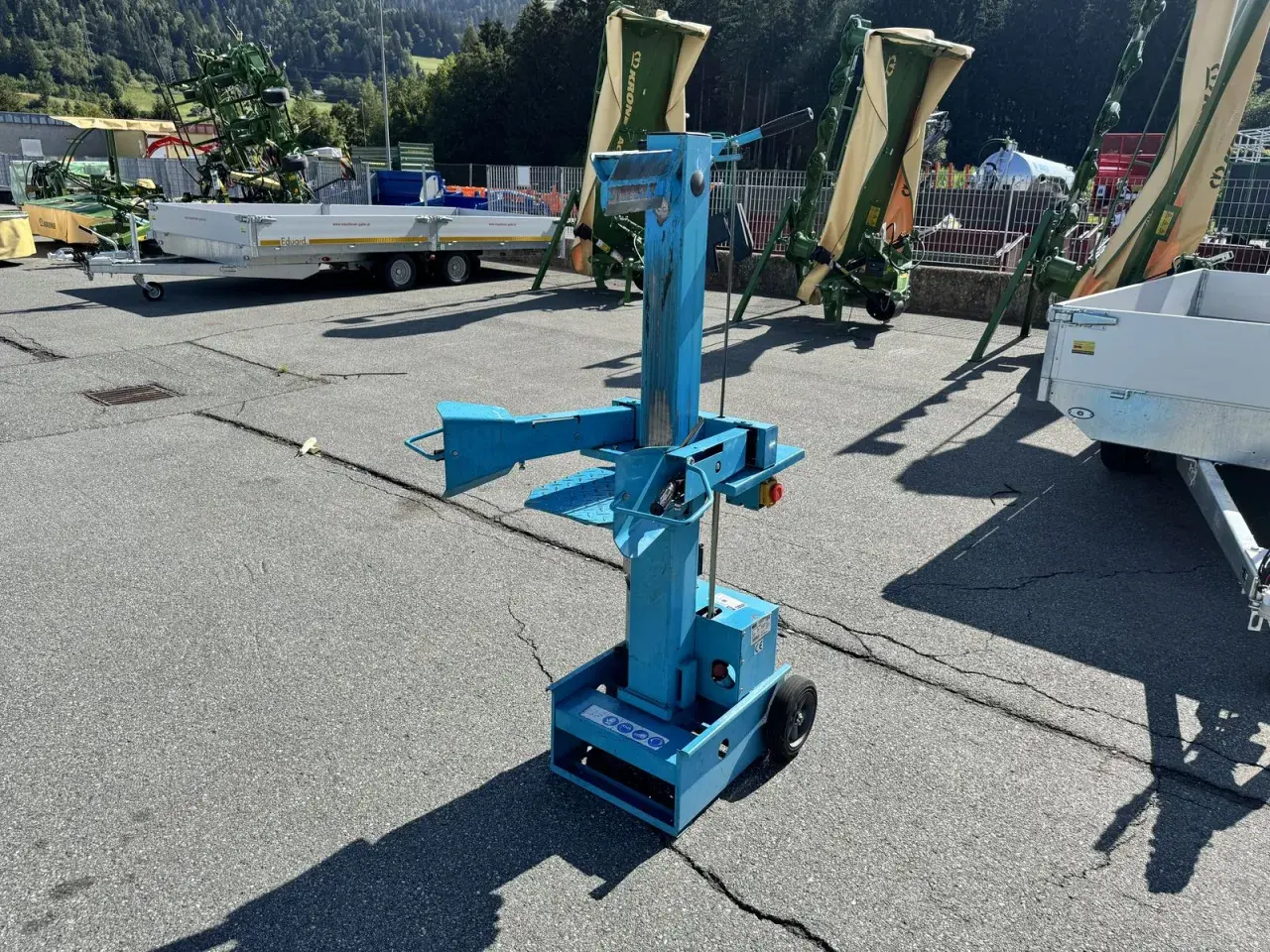 Blauer, gebrauchter Holzspalter mit Rädern steht auf einem Parkplatz. Im Hintergrund landwirtschaftliche Maschinen und Anhänger.