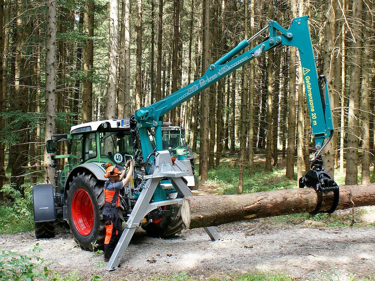 Ein Mann bedient den Kran eines Traktors im Wald. Das Schwenkwerk des Krans greift einen Baumstamm.