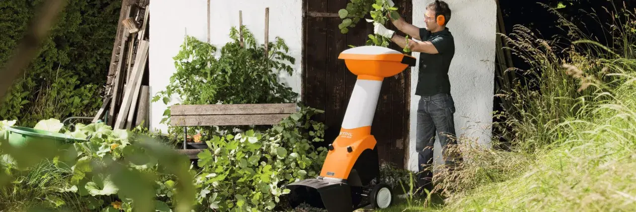 Ein Mann mit Schutzbrille und Gehörschutz bedient einen orange-weißen Holzhacker. Die Zapfwelle des Geräts ist bereit für den Einsatz im Garten.