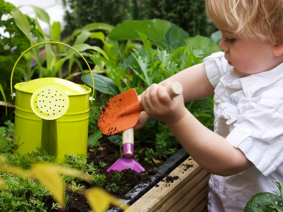 Kleines Kind mit Schaufel im Garten, lernt, wie man ein kinder hochbeet selber baut. Grüne Gießkanne daneben.
