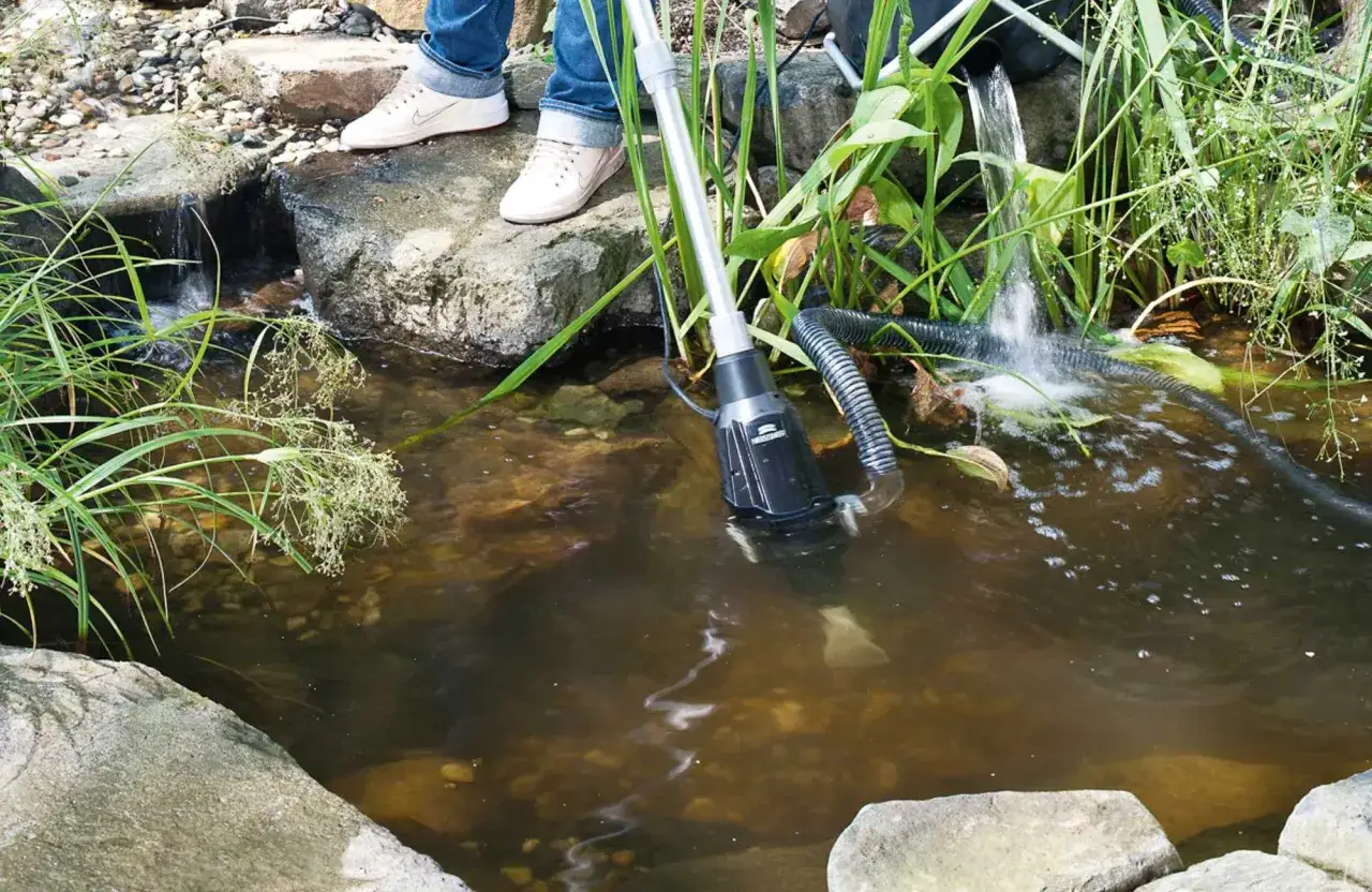 Ein Teichsauger reinigt den naturteich garten. Wasser plätschert über Steine, während eine Person mit dem Gerät im trüben Wasser hantiert.