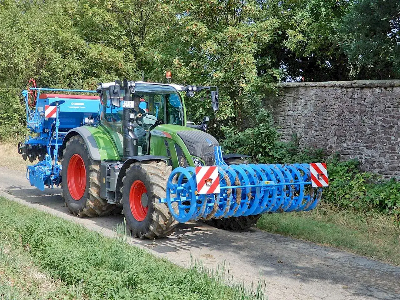 Grüner Traktor mit angehängter Sämaschine und Bodenwalze. Die Bodenwalze hat eine Warnafel halterung mit rot-weißen Warnstreifen.