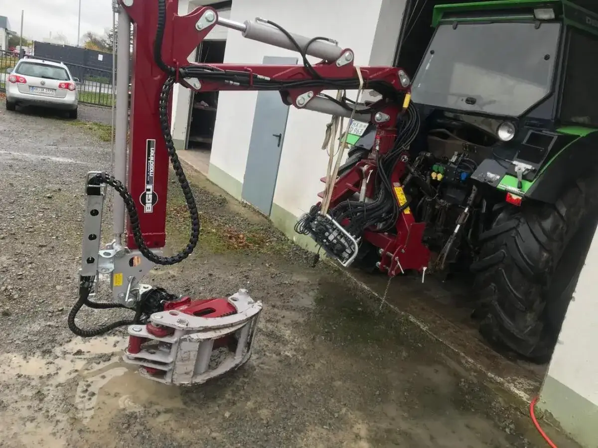 Ein roter BGU Rückezange-Greifer liegt im Matsch, bereit für den Einsatz mit dem danebenstehenden Traktor.