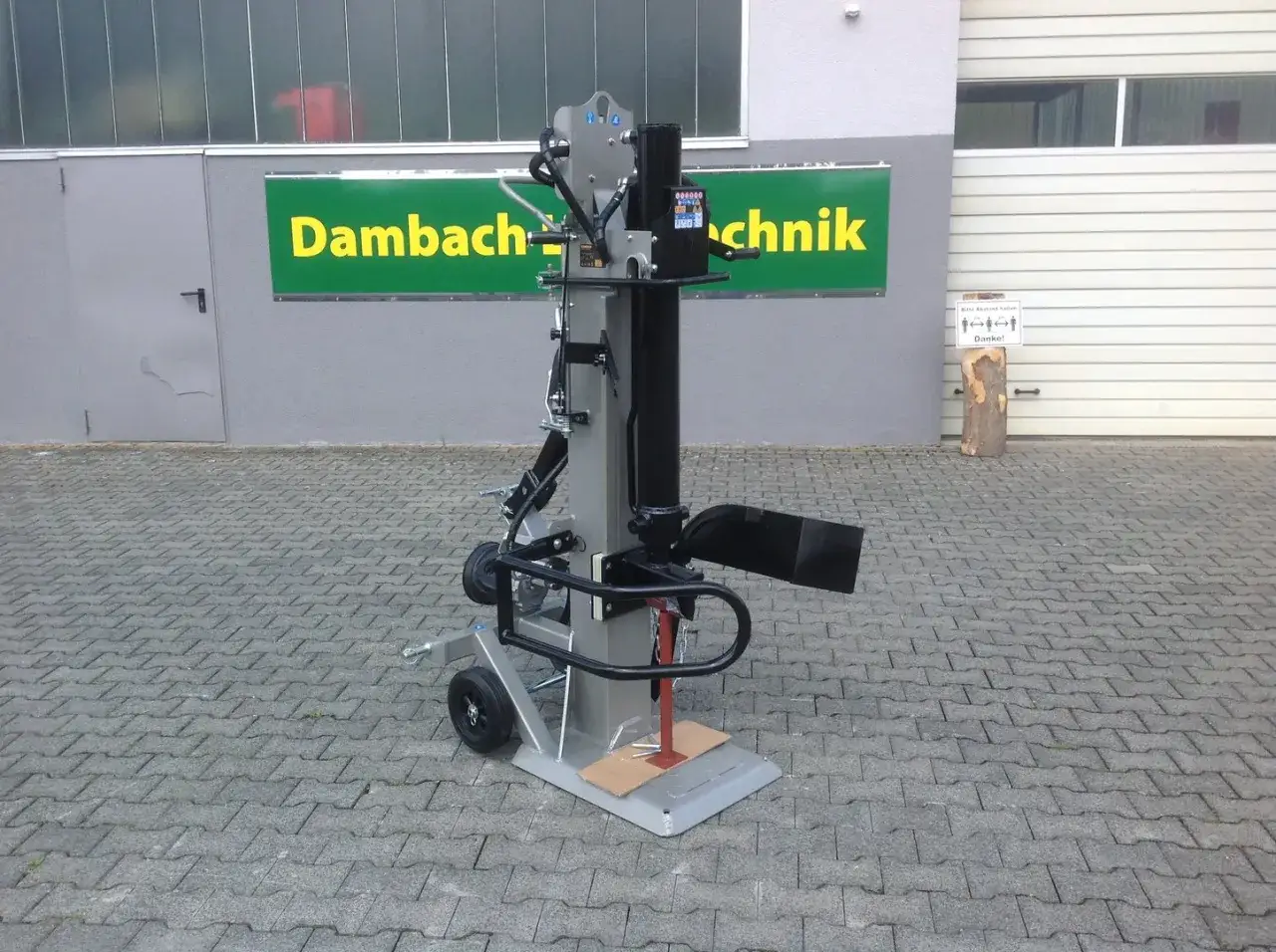 Ein robuster Holzspalter, ca. 1m hoch, steht auf einem gepflasterten Hof vor einem Gebäude mit der Aufschrift