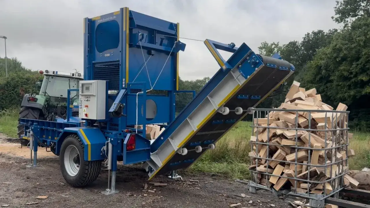 Ein blauer Förderband für Holz transportiert gespaltene Holzscheite in einen Gitterkorb. Ein Traktor steht daneben.