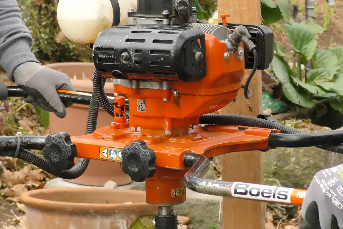 Eine orangefarbene Erdbohrer Maschine mit schwarzen Griffen und einem Motor. Eine Hand in einem grauen Handschuh hält einen der Griffe.