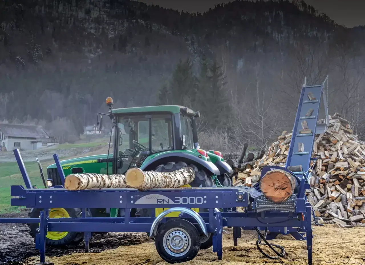 Ein Traktor mit einem Holzspalter Tajfun RCA 400 bereitet Holz vor. Ein großer Holzstapel liegt im Hintergrund.