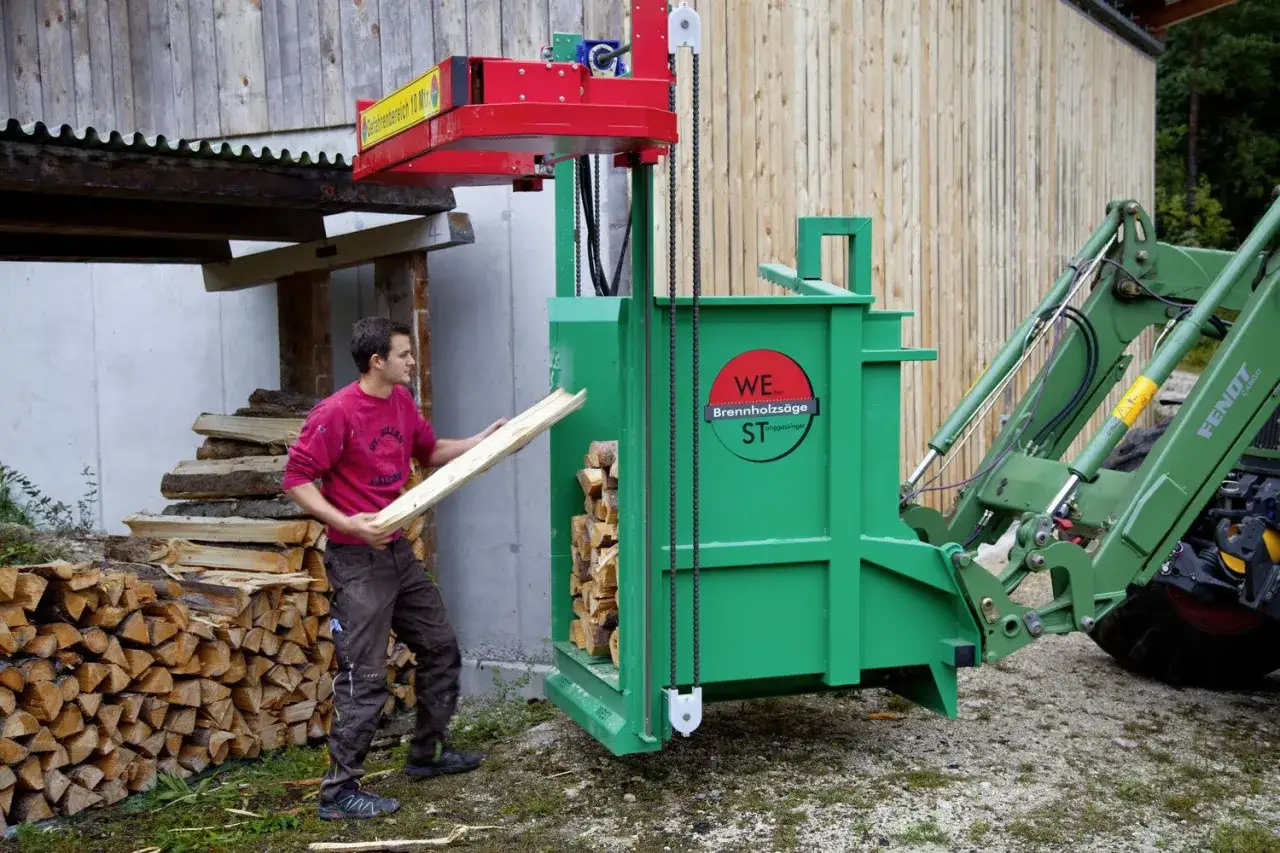 Mann belädt ein grünes Bündelgerät mit Holzscheiten. Ein Traktor steht bereit.