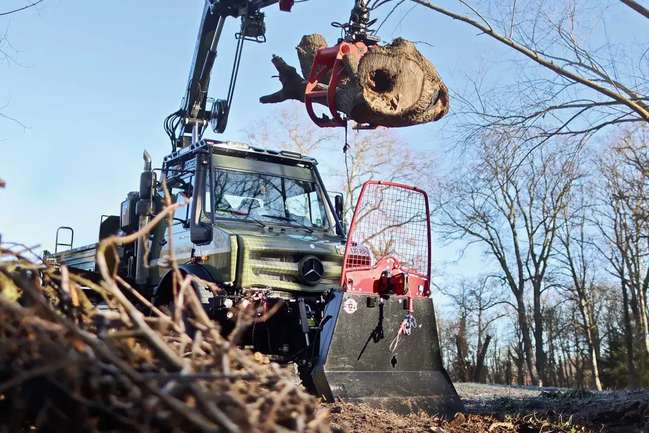 Grüner Ladekran hebt Baumstamm mit einer roten Chokerkette. Schneeschild am Unimog.