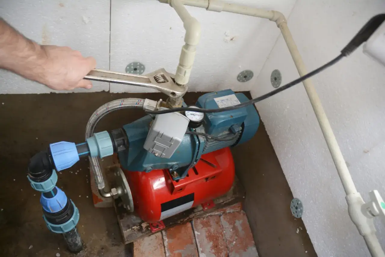 Hand repariert ein Hauswasserwerk für den Garten. Die Pumpe mit rotem Tank und blauem Gehäuse wird mit einem Schraubenschlüssel angeschlossen.