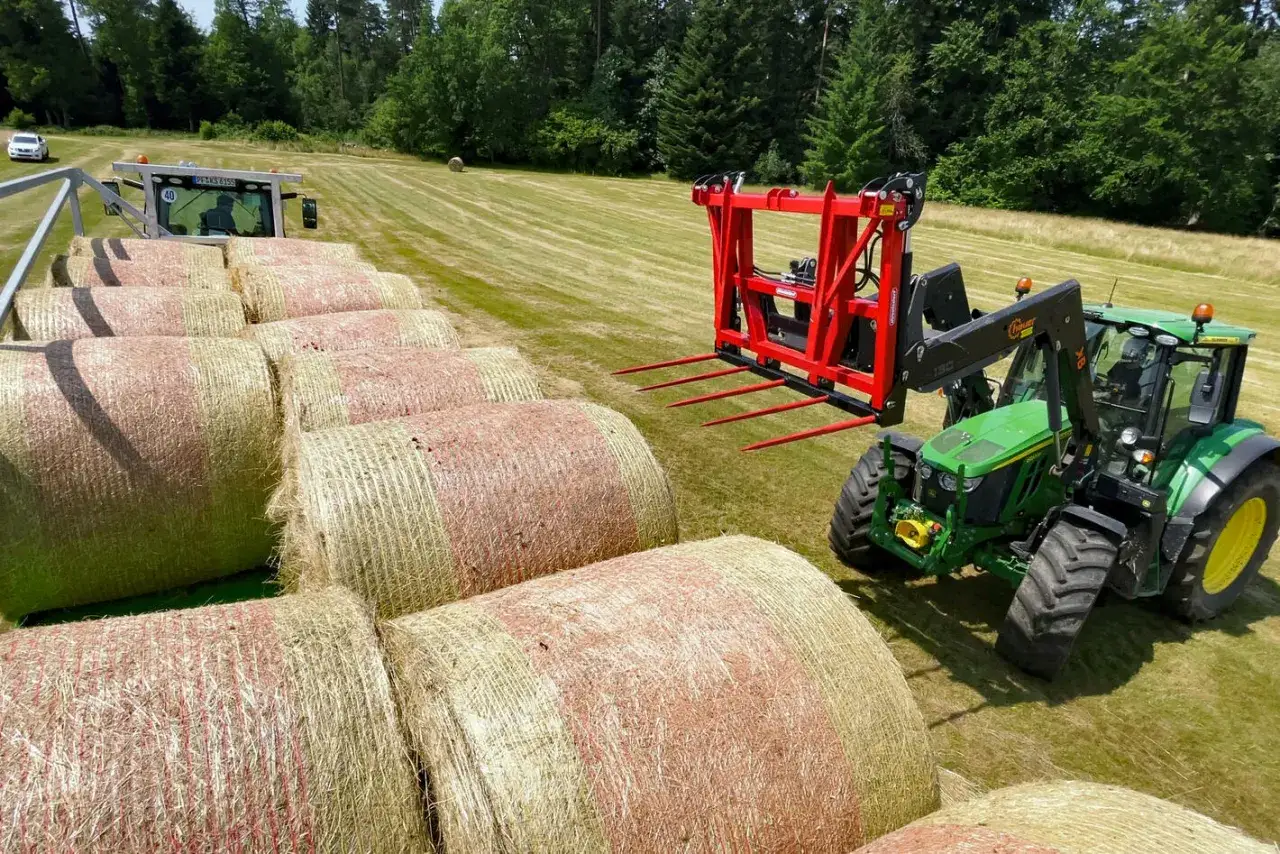 Grüner Traktor mit roter Ballentransportgabel hebt Strohballen auf einem Feld.