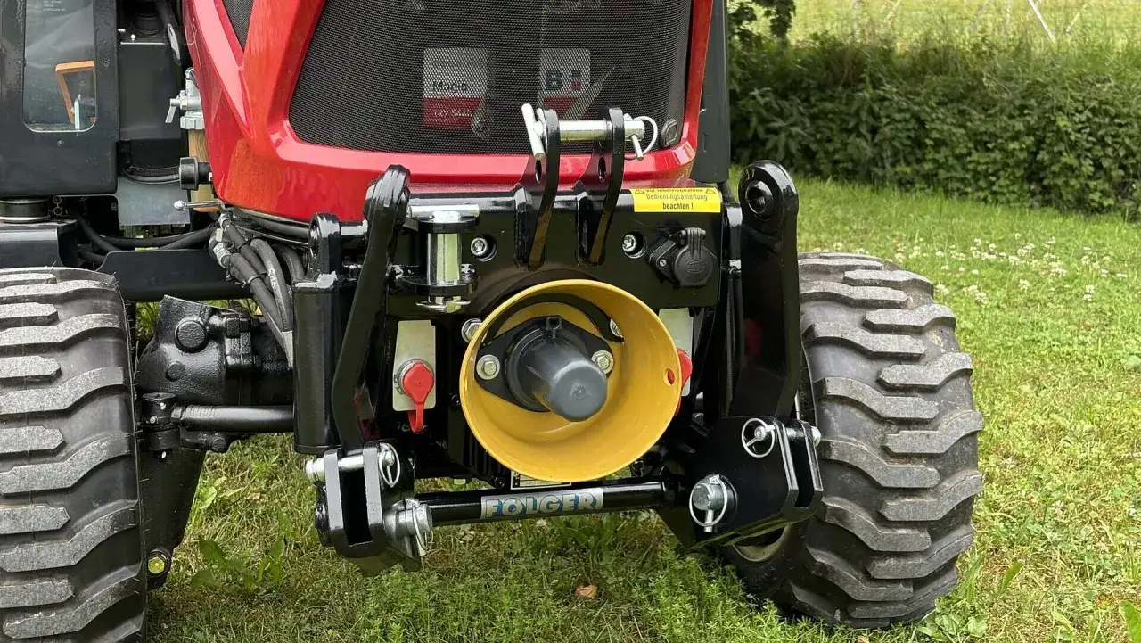 Traktor mit gelbem, weistem dreieckförmigem Anbaugerät und schwarzer Folger-Deichsel.