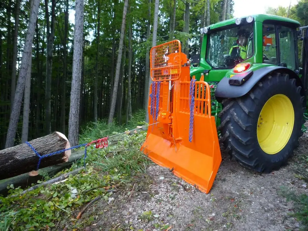 Ein kleiner Forsttraktor mit orangefarbenem Seilwinden-Anbau zieht einen Baumstamm im Wald.