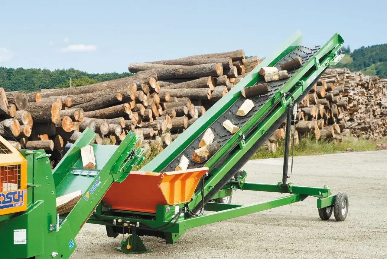 Grüne mobile Förderbänder transportieren Holzscheite vor einem großen Stapel Baumstämme.
