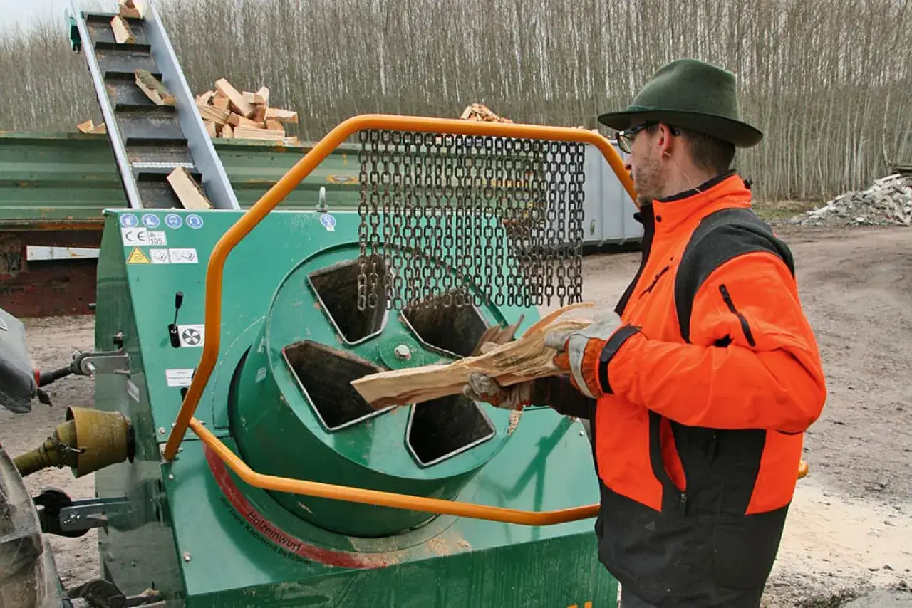 Mann mit orangefarbener Jacke und Hut füttert Holzscheite in eine grüne Wippsäge, die Holz zerkleinert.