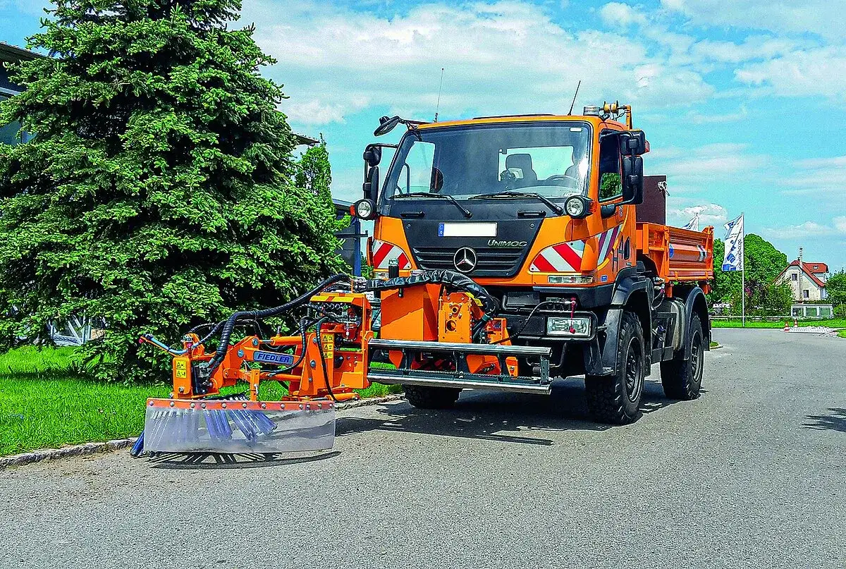 Ein Unimog mit einer Fiedler-Wildkrautbürste auf einer Straße. Die Bürste ist vorne angebracht und bereit, Unkraut zu entfernen.