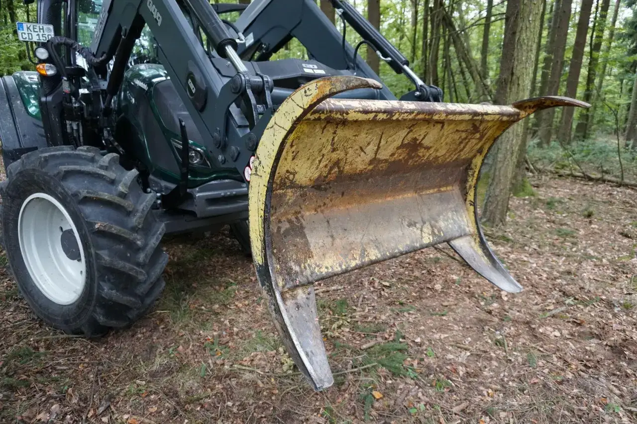 Ein grüner Traktor mit einem gelben, abgenutzten Frontpolterschild steht im Wald.
