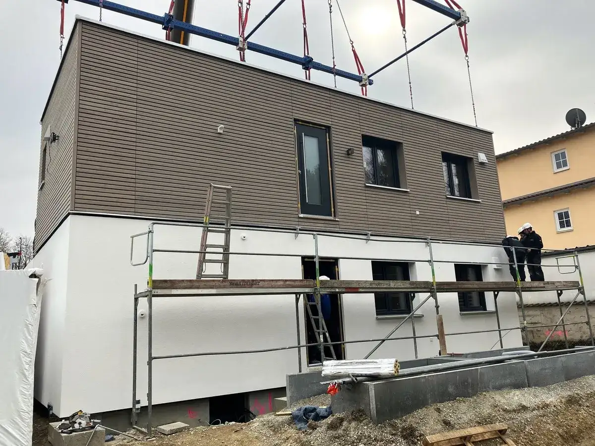 Ein Kran hebt ein Holzhaus-Modul in einem Wohngebiet. Bauarbeiter auf Gerüsten arbeiten am Haus.