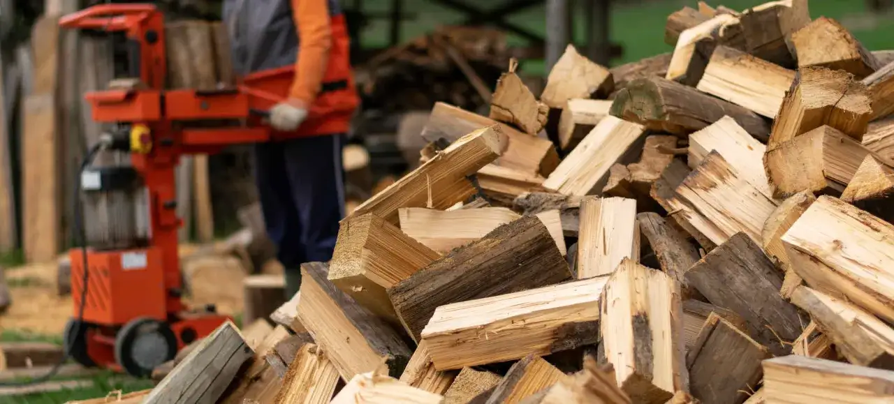 Ein Mann benutzt einen Holzspalter, um einen Baumstamm zu spalten. Ein großer Haufen gespaltenen Holzes liegt im Vordergrund.