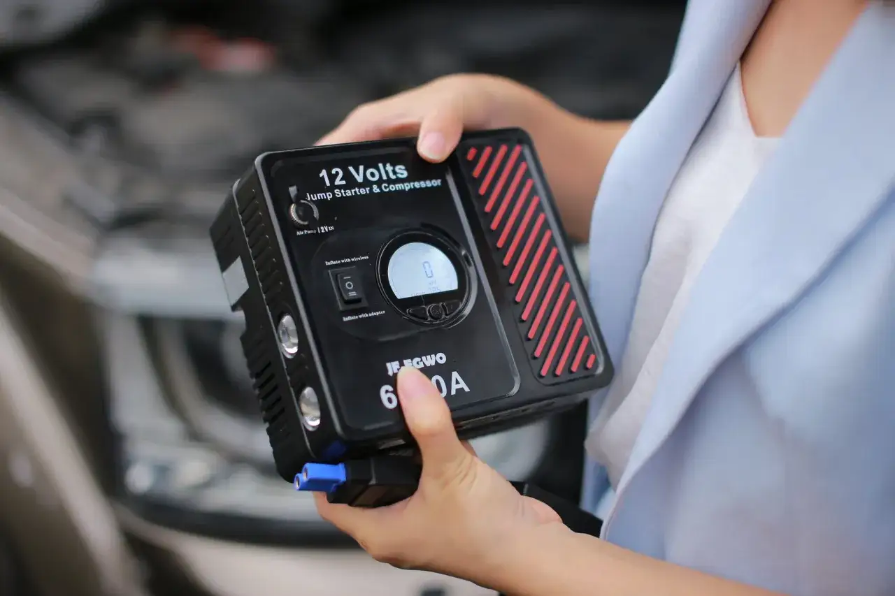 Hände halten einen 12-Volt-Batteriestarter und Kompressor. Das Gerät ist bereit, bei Bedarf Starthilfe zu geben oder Reifen aufzupumpen.