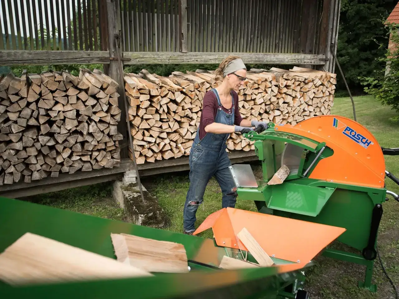 Frau bedient eine rolltischsäge, um Holzscheite zu schneiden. Im Hintergrund sind Holzstapel zu sehen.