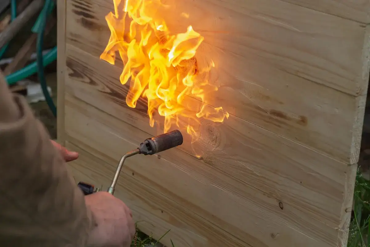 Flammen lodern auf hellem Holz, das sehr alt ist und schlecht brennt. Ein Mann hält einen Brenner.