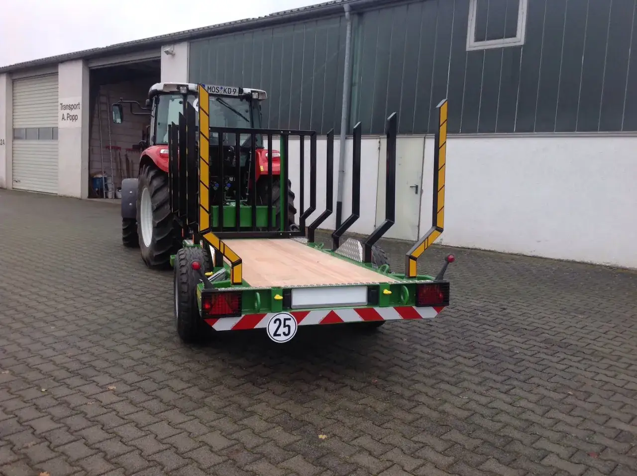 Ein roter Traktor mit einem neuen meterholzwagen mit kran steht vor einer Halle.