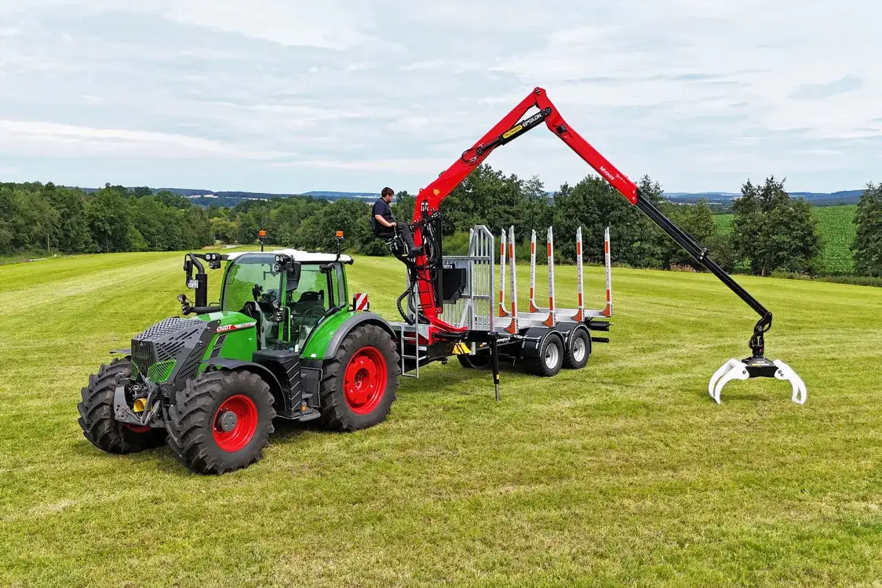 Grüner Traktor mit angekoppeltem Anhänger und einem roten Ladekran, bereit für den Holzeinsatz im Wald.