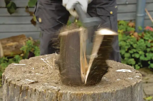 Ein Mann spaltet mit einem Keil einen Baumstamm. Holzsplitter fliegen durch die Luft.
